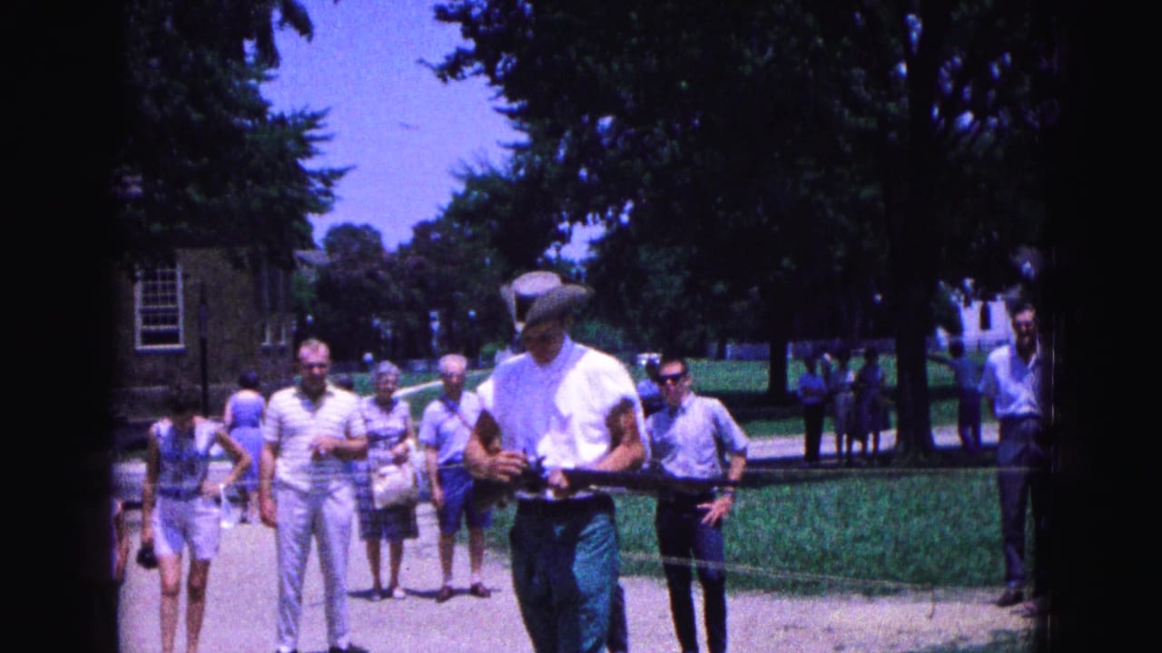 Costumed man with a long gun walks near tourists on a sunny path.
