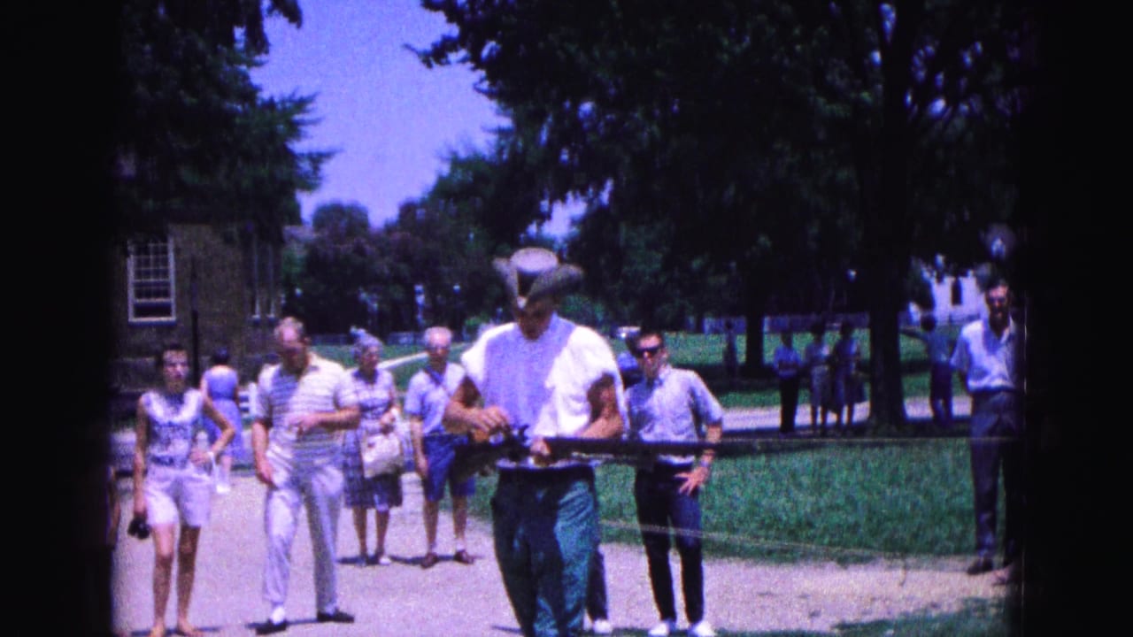 A Williamsburg visitor scene shows tourists in 1960s clothing watching a costumed man carry a long gun beside a rope barrier.