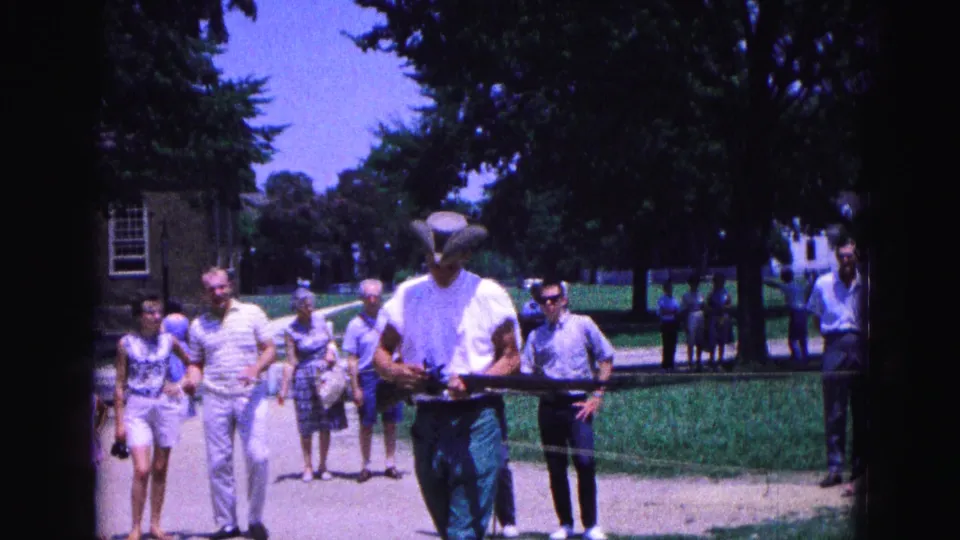 The 1964 Williamsburg footage shows the costumed man holding a long gun in the foreground with tourists and trees behind him.
