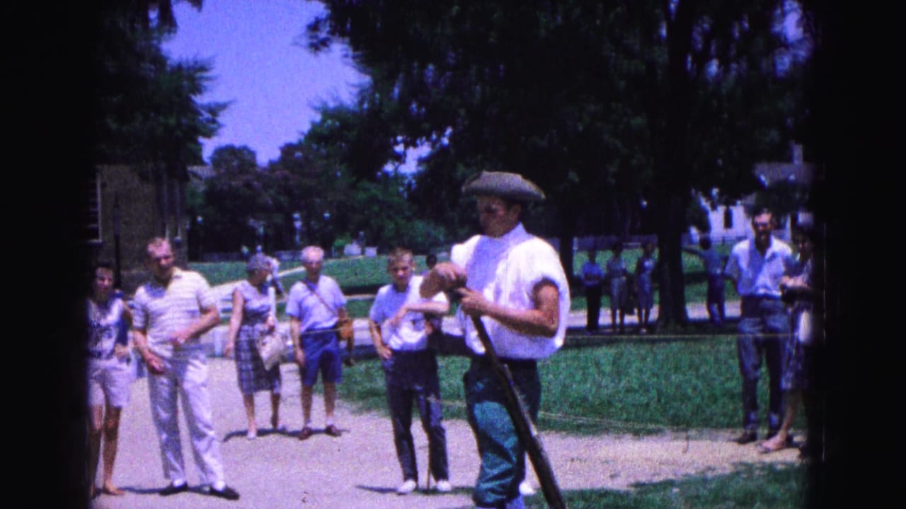 Costumed interpreter holding a musket while tourists watch outdoors.