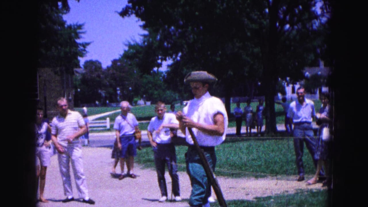 A Williamsburg reenactor in period-style dress stands with a musket while visitors watch from a shaded outdoor area.