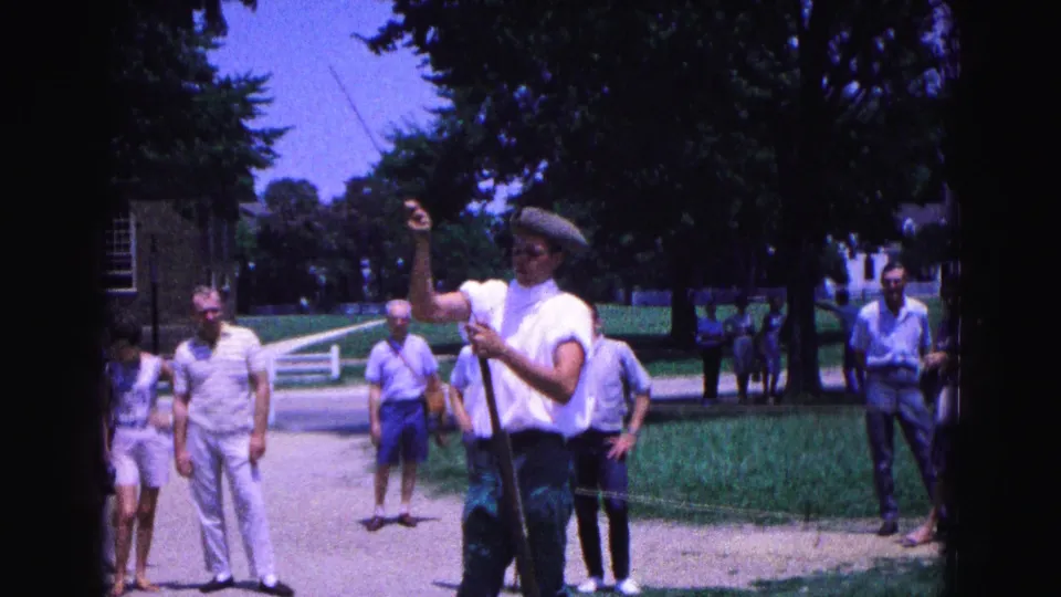 The interpreter gestures upward during the Williamsburg musket demonstration as tourists stand nearby on the walkway.