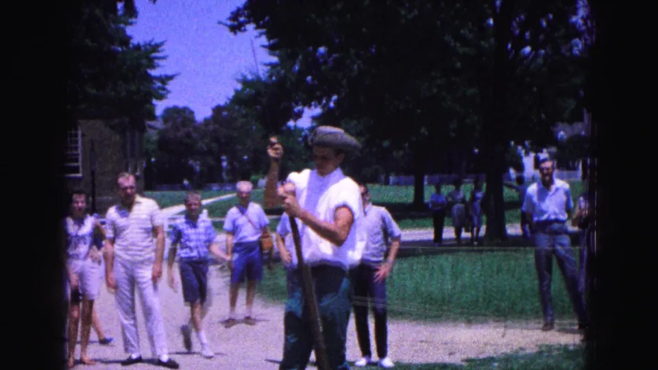 A costumed Williamsburg interpreter holds the musket upright with tourists and trees visible behind him.