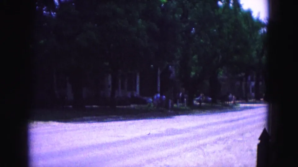 1964 Williamsburg home-movie frame showing a quiet tree-lined road with shaded residential frontage and dark film gate edges.