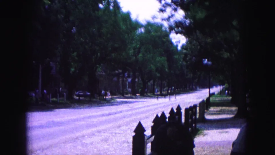 Williamsburg, Virginia 1964 frame showing a bright road edge, shaded trees, fence posts, parked cars, and small figures walking in the distance.