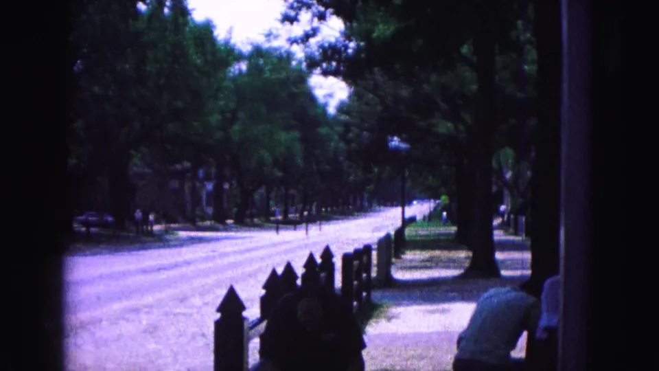 1964 Williamsburg roadside frame with a shaded fence line, large tree trunk, parked cars, distant pedestrians, and partial foreground figures at right.