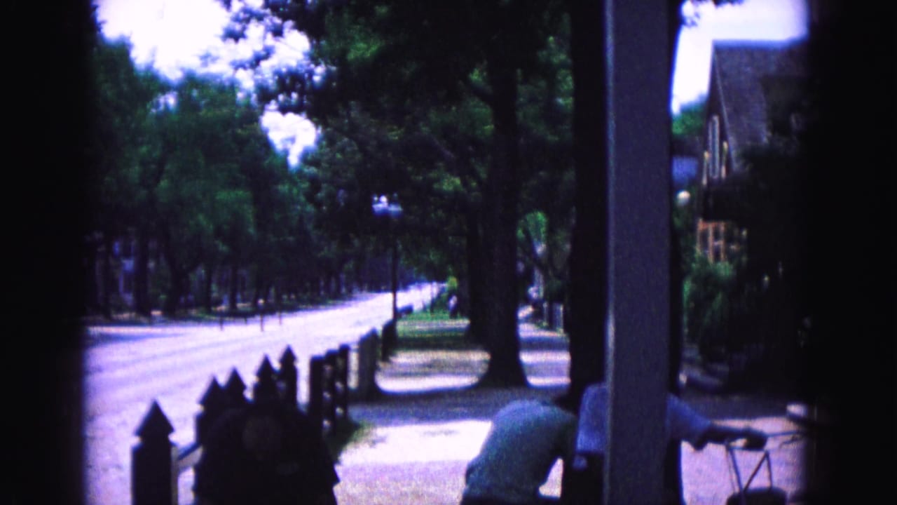 Williamsburg home-movie frame showing a shaded porch, quiet road, low fence, and a person in a light shirt at the edge.