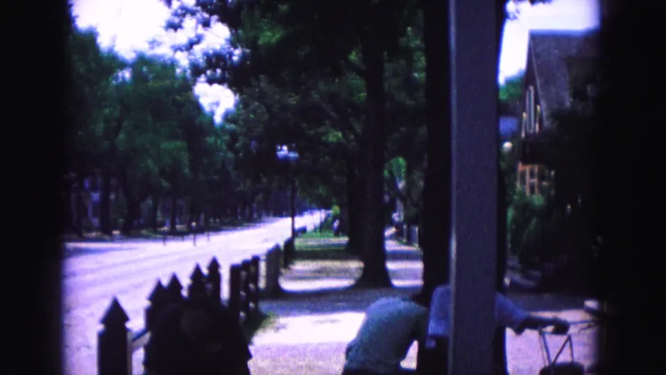 Williamsburg home-movie frame showing a shaded porch, quiet road, low fence, and a person in a light shirt at the edge.