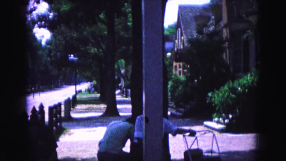 1964 Williamsburg footage pans from the street toward a porch-side view with shrubs, house facade, and a person bending near the foreground.