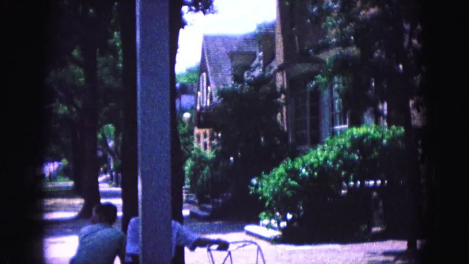 Williamsburg residential exterior with porch columns, dense shrubs, shaded windows, and a person partly visible in 1964 home-movie footage.