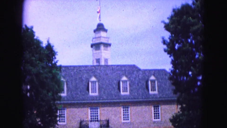 1964 Williamsburg, Virginia home-movie frame showing a brick historic-style building with dormers, cupola, and trees at the edge of the view.