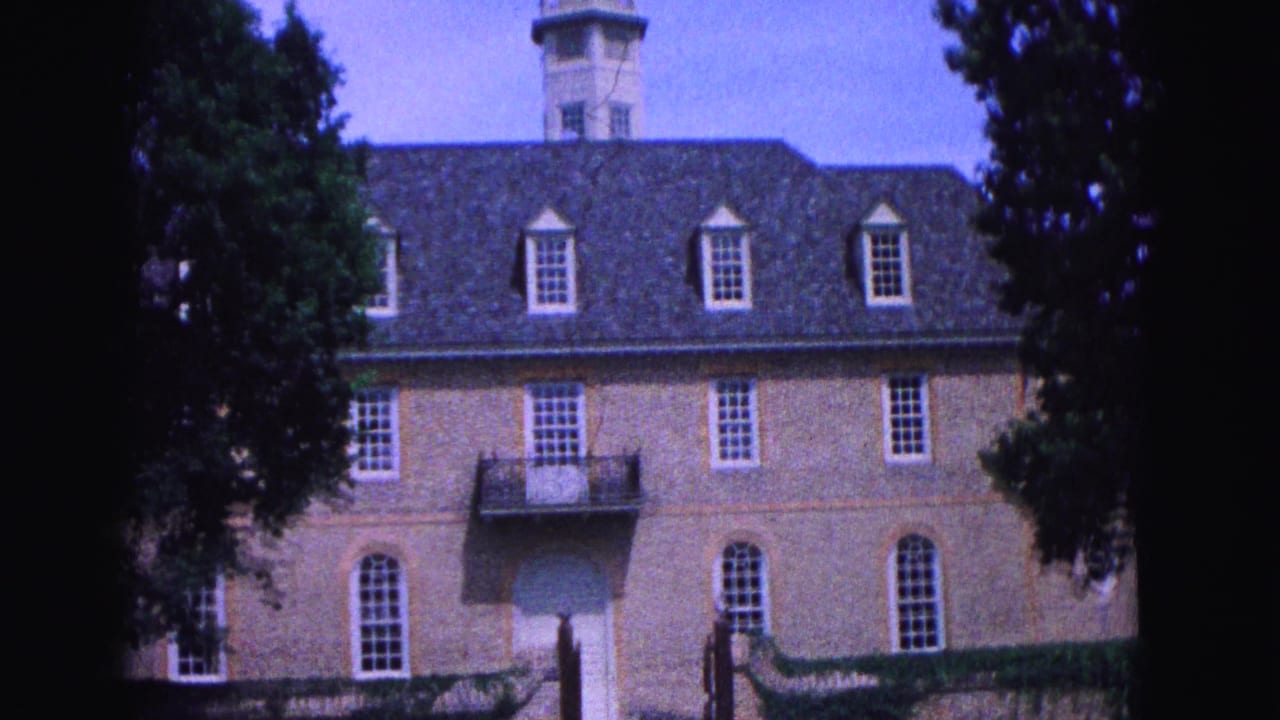 1964 Williamsburg, Virginia frame of a large brick building with dormer windows, a cupola, balcony, and surrounding trees.