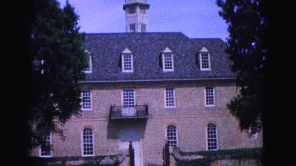 1964 Williamsburg, Virginia frame of a large brick building with dormer windows, a cupola, balcony, and surrounding trees.