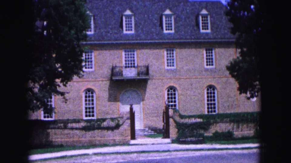 1964 Williamsburg, Virginia view of a brick building entrance with dormers, arched windows, trees, a stone wall, and a walkway.