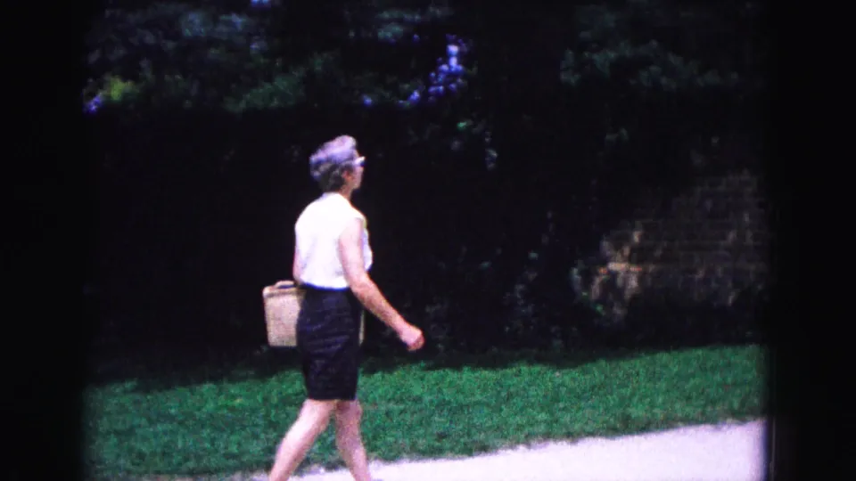 1964 Williamsburg, Virginia home-movie frame showing an elderly woman in a white top and dark skirt carrying a basket along a path.