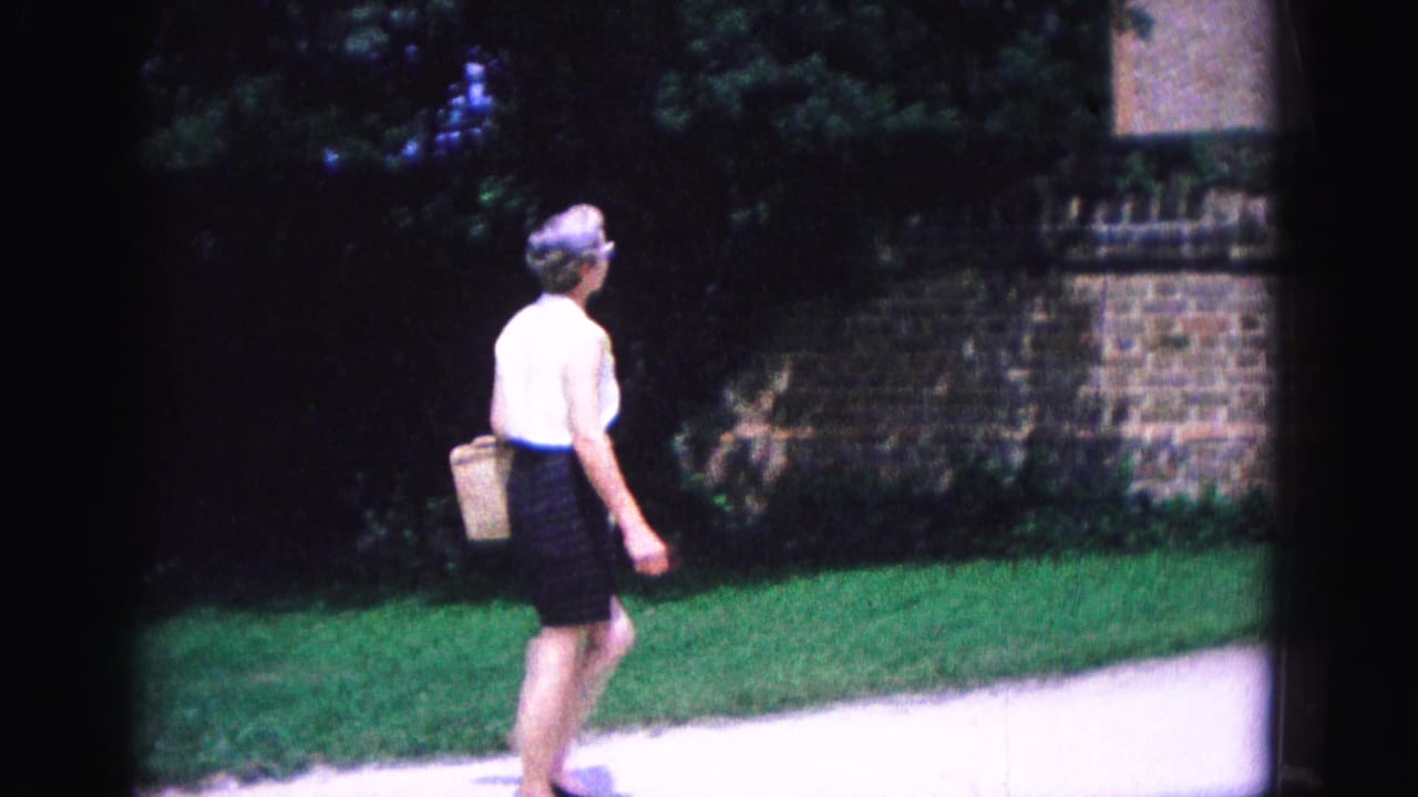 Woman in a white sleeveless top and dark skirt walking beside a brick wall and grass.