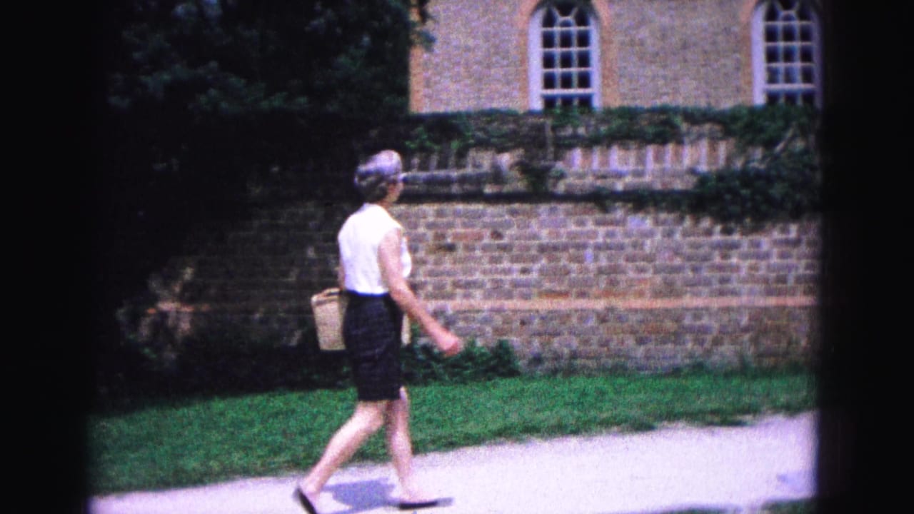 A woman with glasses and a woven basket walks past a brick wall and arched windows in Williamsburg, Virginia, in 1964.