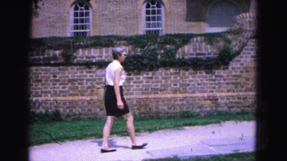 The 1964 Williamsburg footage shows the woman continuing beside an ivy-covered brick wall with arched windows above.