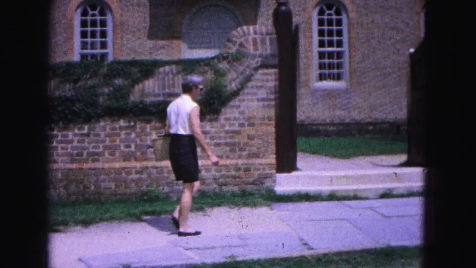At the end of the shot, the woman walks toward a gate or doorway set into the brick streetscape in 1964 Williamsburg.