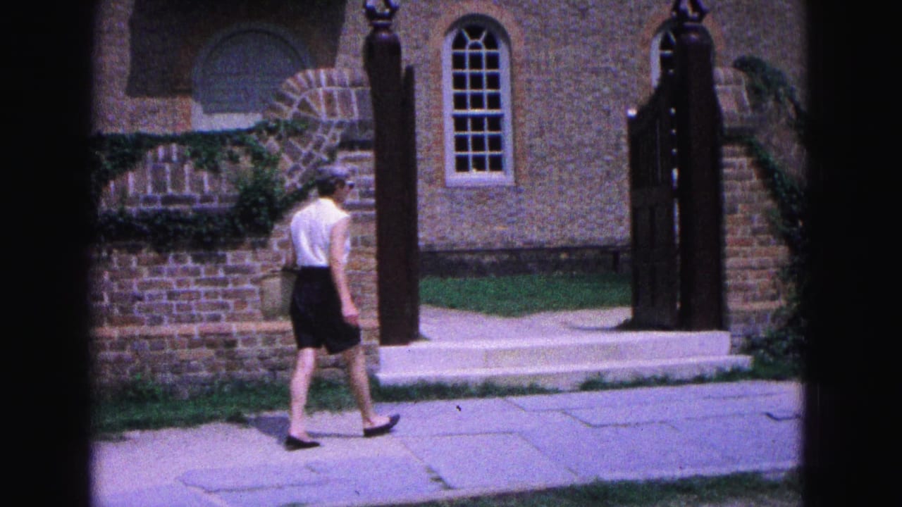 In Williamsburg, Virginia, a woman walks past brick walls, arched windows, and black gateposts in 1964 home-movie footage.