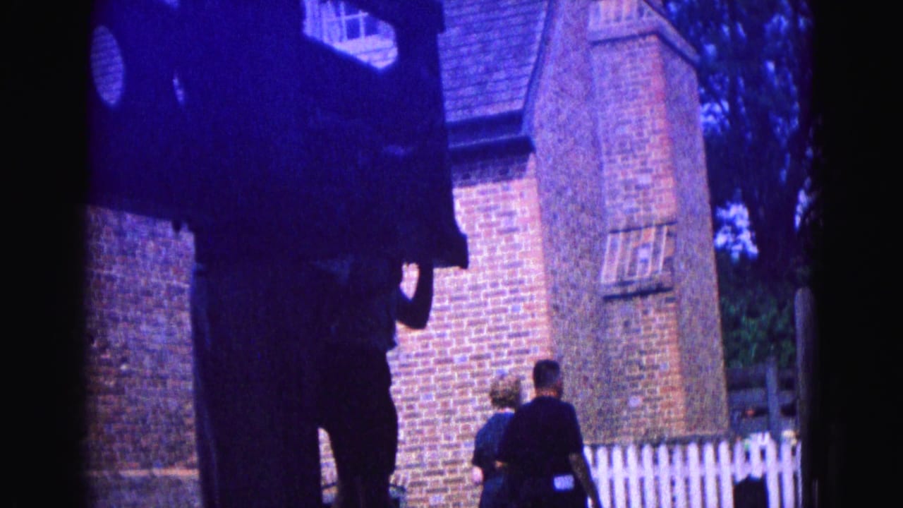 Visitors walk beside a brick building and white fence, partly blocked by a dark foreground object.