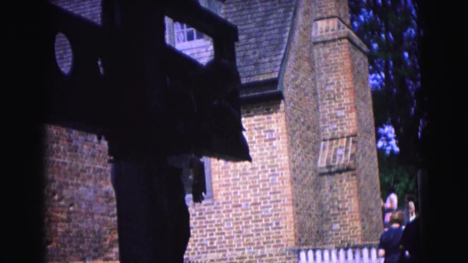 Home-movie frame of a Williamsburg brick structure, partially obscured by a dark object, with visitors visible near the right edge.