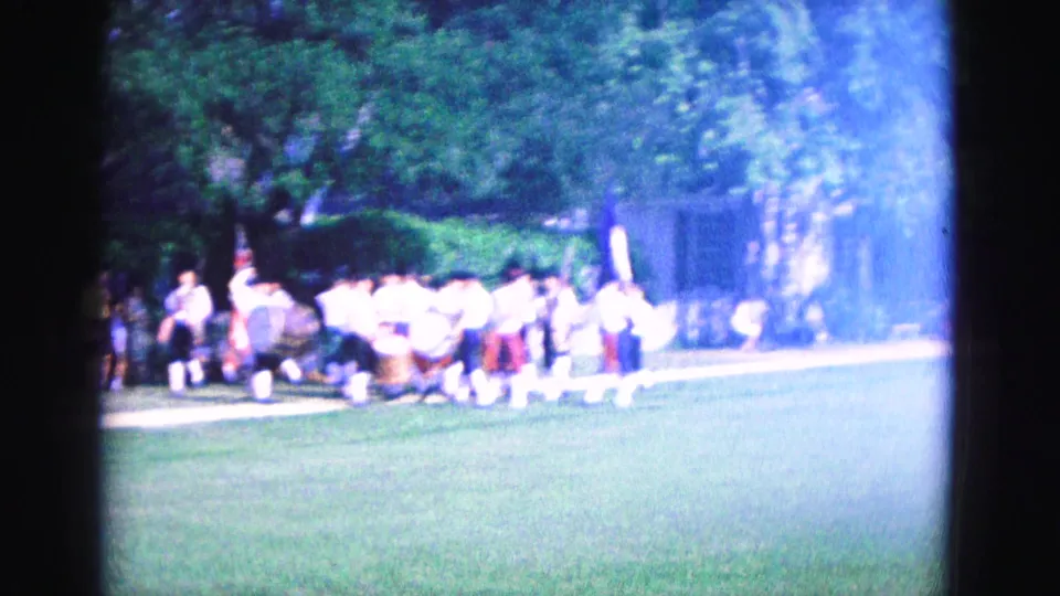 1964 Williamsburg frame showing a distant uniformed band or ceremonial group with flags on a lawn, filmed with bright exposure shift.