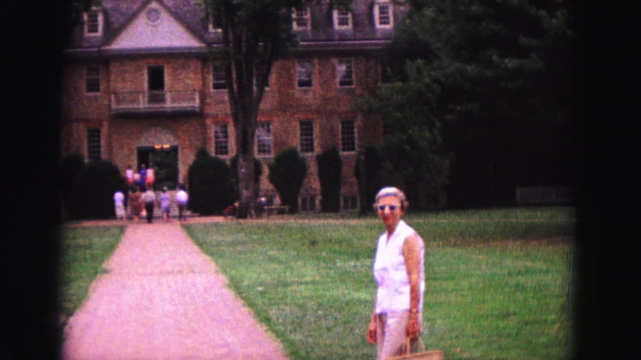 Woman in white standing on a lawn before a large brick building with visitors near the entrance.