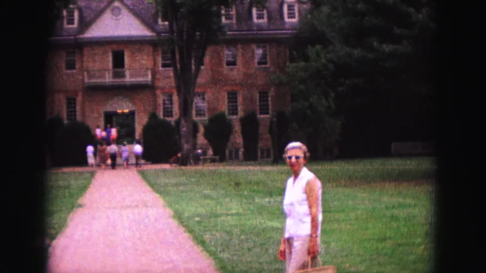 In 1964 Williamsburg, a woman in sunglasses stands on grass beside a path leading toward a large brick building with visitors at the doorway.