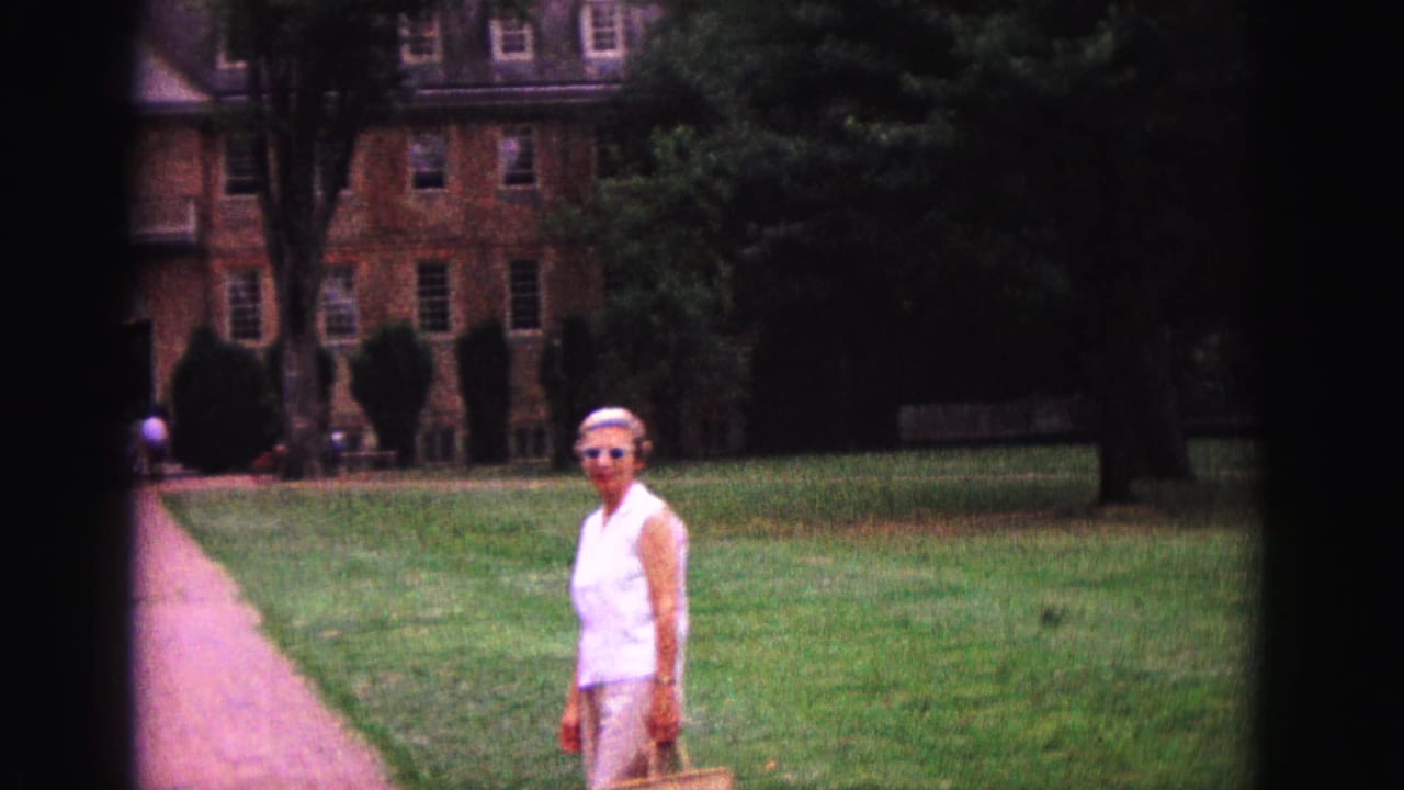 A 1964 Williamsburg home-movie frame shows a woman standing on the lawn beside a paved path, with a brick building and trees in the background.