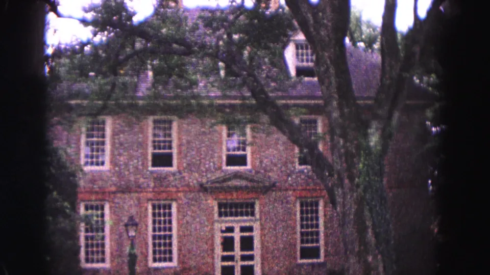1964 Williamsburg, Virginia 8mm frame showing a brick multi-story building with white-trimmed windows behind large tree trunks and leafy branches.