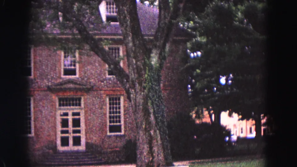 1964 Williamsburg, Virginia home-movie frame with a closer view of a brick building entrance, tall windows, tree bark, and heavy shade at the frame edge.