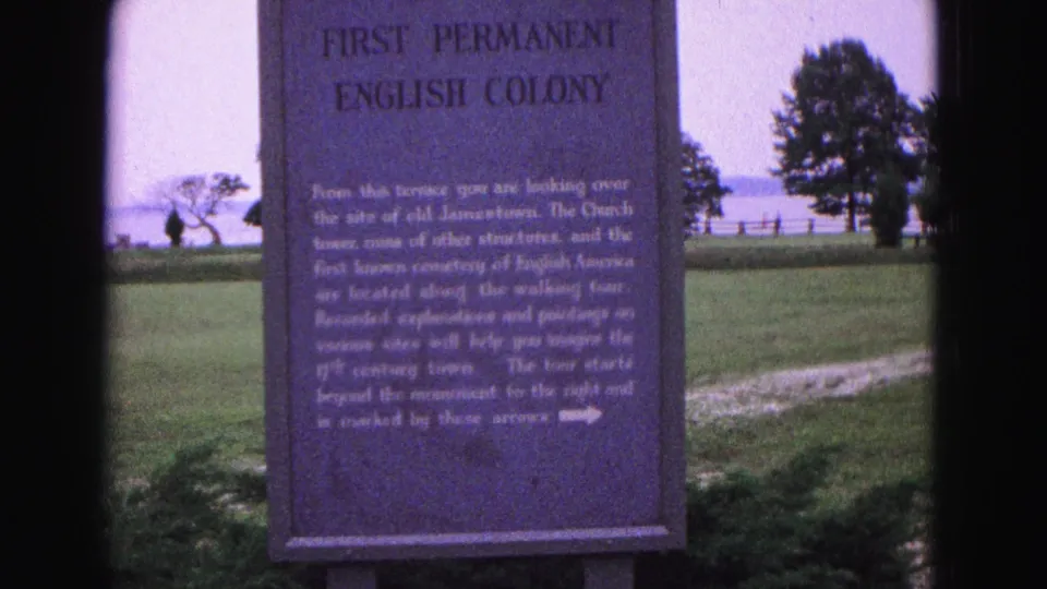 1964 Williamsburg frame showing the First Permanent English Colony marker with a lawn, trees, fence, distant water, and small visitors in the background.