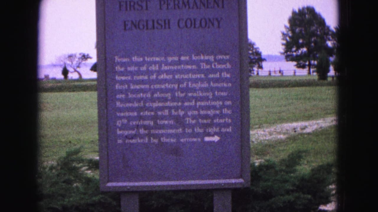 Historical marker beside a lawn with water and trees in the background.