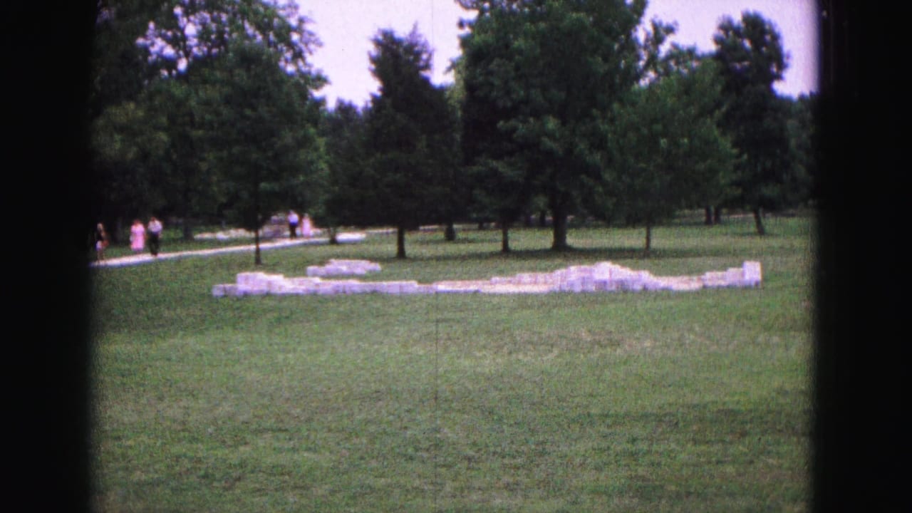 Grassy park with low white foundation ruins, trees, path, and distant visitors.