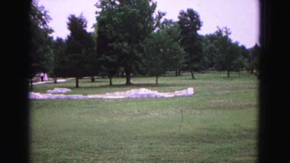 1964 8mm frame of Jamestown-area ruins on grass, with trees and small distant visitors along the path.
