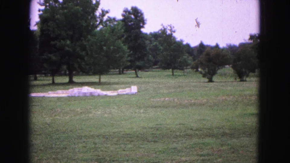 1964 Williamsburg-area 8mm frame showing low foundation remains in an open grassy historic landscape with trees behind.