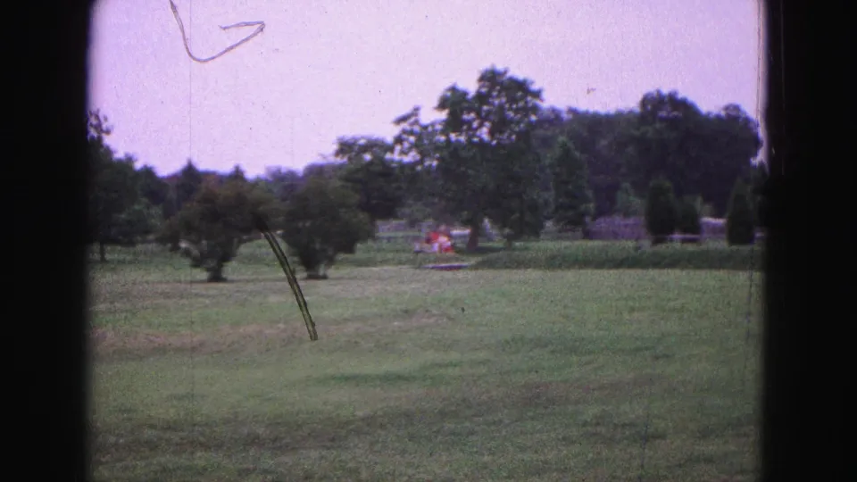 1964 Williamsburg 8mm frame showing a landscaped grassy area with trees, shrubs, distant indistinct people or flowers, and film wear.