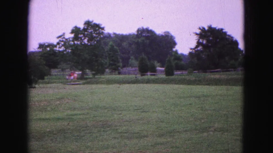 Final 1964 Williamsburg frame with open lawn, mature trees, shrubs, a low boundary, and tiny indistinct figures or color patches in the distance.