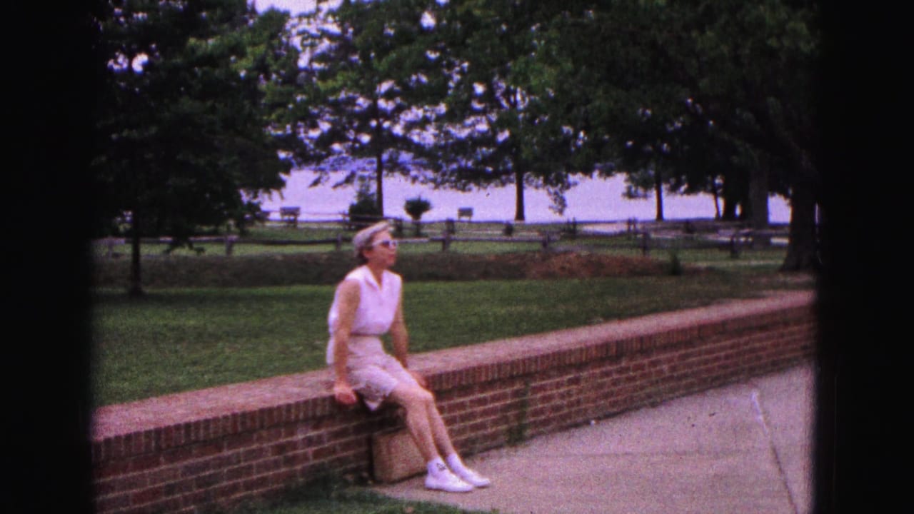 Woman in sunglasses sits on a low brick wall beside grass and trees.