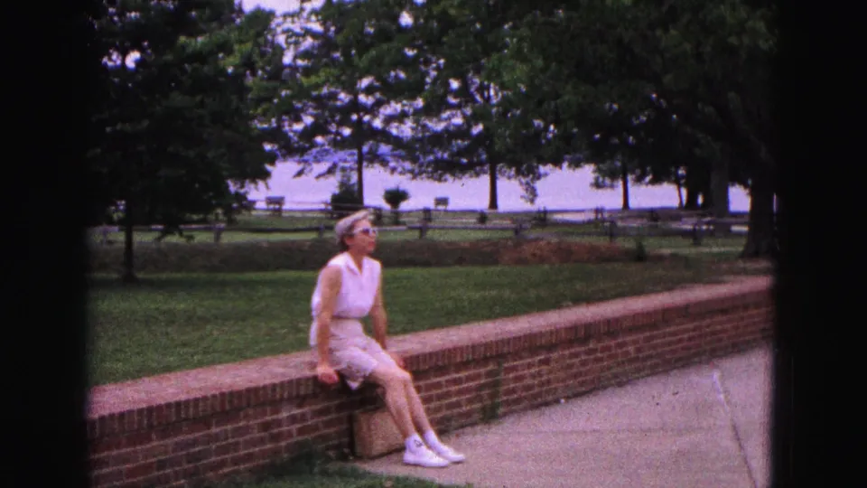 In 1964 Williamsburg, Virginia, a woman in summer clothing sits on a low brick wall near a walkway, grass, trees, fence, and distant water.