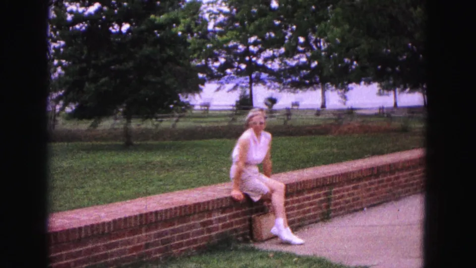 The 1964 Williamsburg home-movie frame shows the woman seated on the brick wall, with soft 8mm color, trees, grass, fence, and water in view.