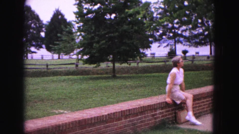 At the end of the clip, the woman sits near the right edge of the Williamsburg park scene, with brick wall, lawn, trees, fence, and water visible.