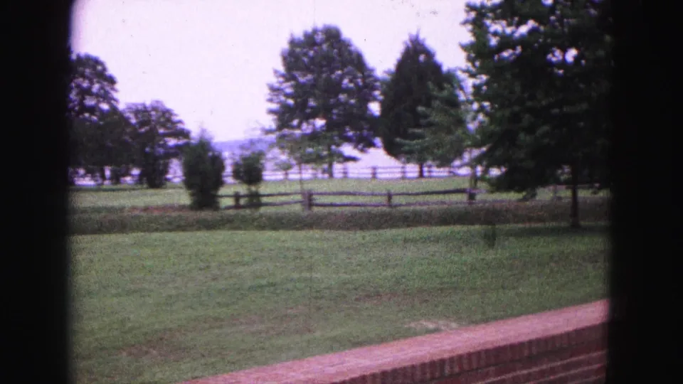 The 1964 Williamsburg view looks across a maintained lawn with a brick wall in front, split-rail fencing, shrubs, and tall trees beyond.