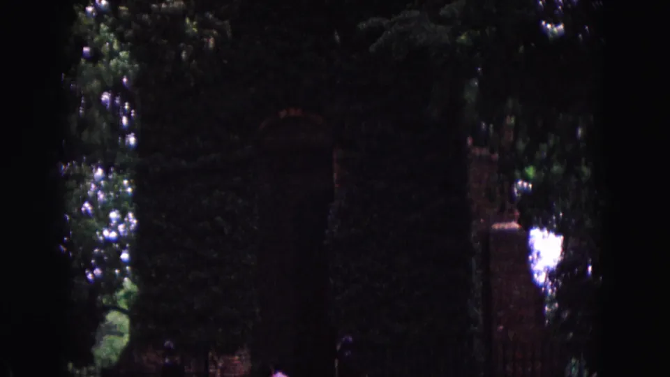 A shadowed Williamsburg brick gateway appears under dense foliage, with ivy-covered walls, iron fencing, and visitors moving near the entrance.