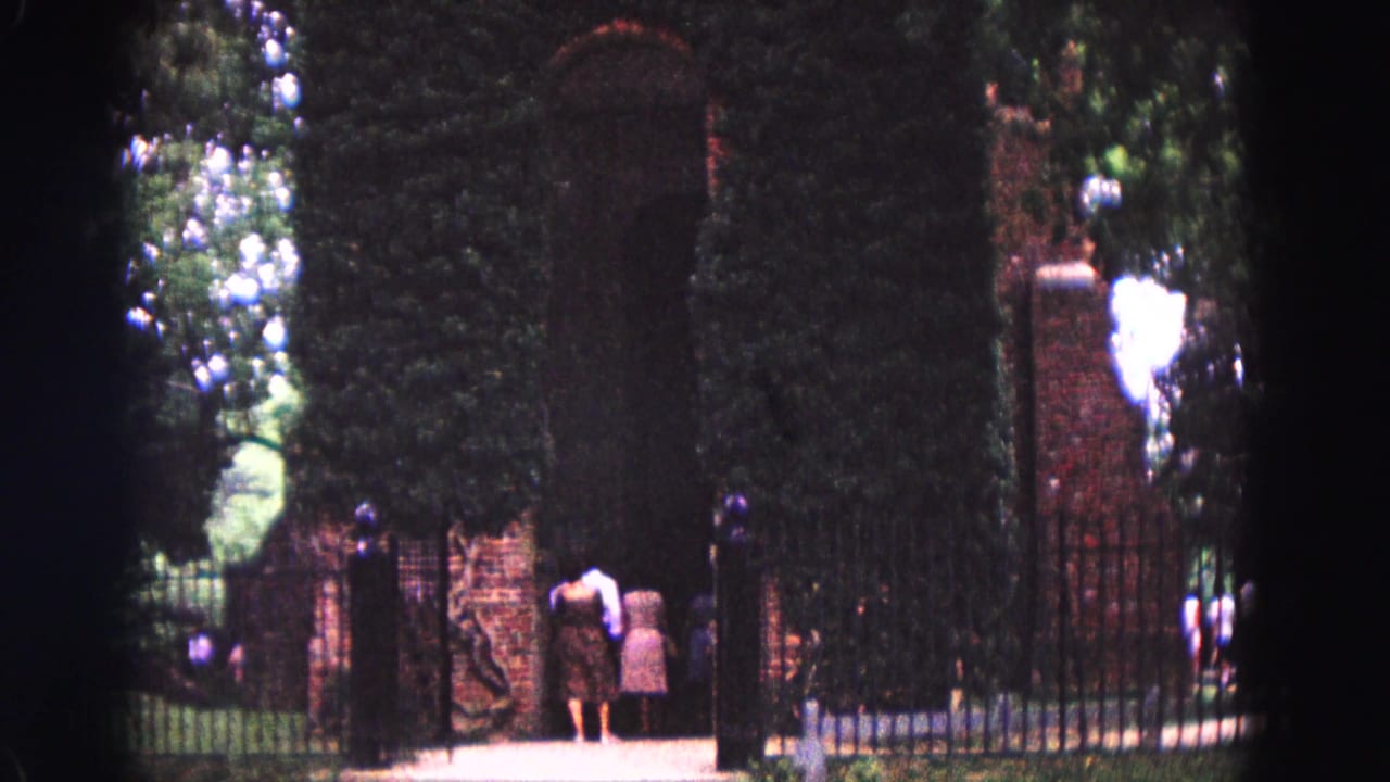 Visitors stand near a brick gate framed by hedges and black iron fencing.