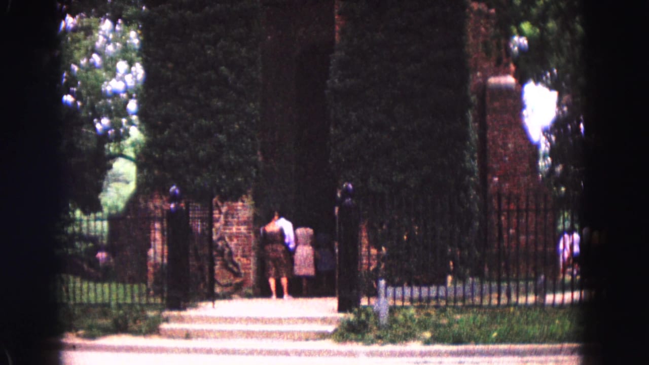 People stand at a gated entrance with brick posts, hedges, and a path beyond.