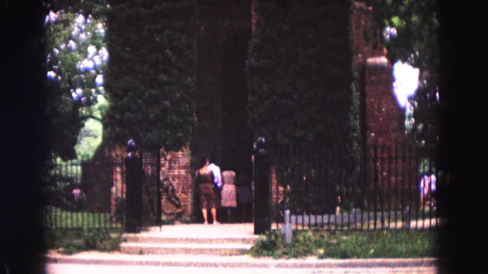 1964 Williamsburg view of people at a historic gated entrance with brick posts, clipped hedges, iron fencing, and greenery beyond.