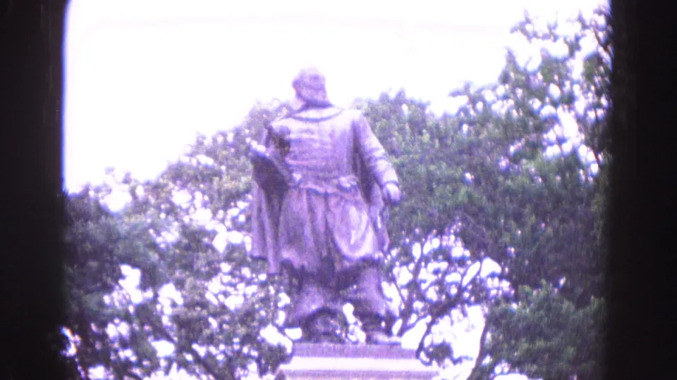 1964 Williamsburg home-movie frame showing a colonial-style statue on a stone pedestal, partly overexposed against trees and sky.