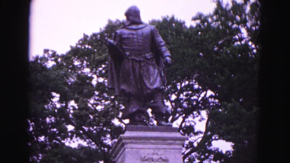 1964 Williamsburg frame showing a colonial-style figure statue on a pedestal, with leafy trees filling the background.